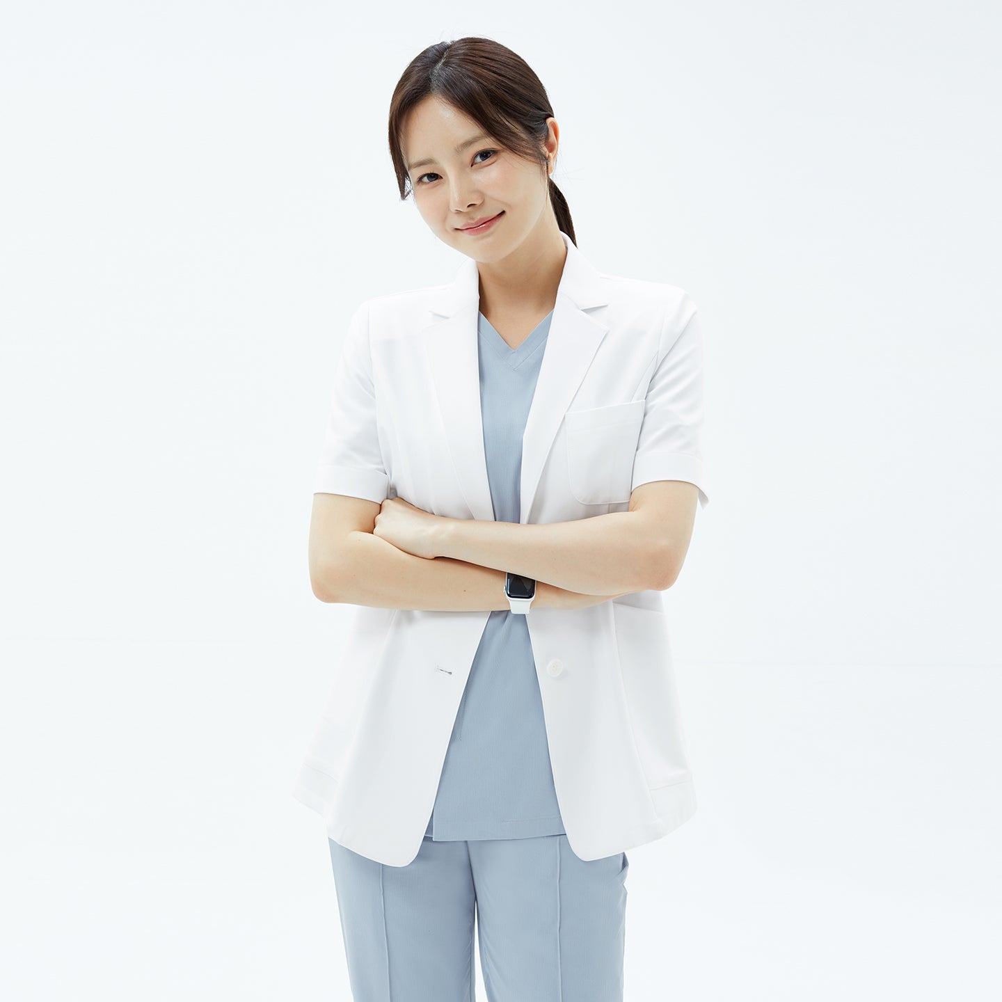 The wearer stands smiling with arms crossed in the TUL Women’s LCW-15 Lightweight Short Sleeve Lab Coat – White, showcasing its patch pockets and lightweight fabric.
