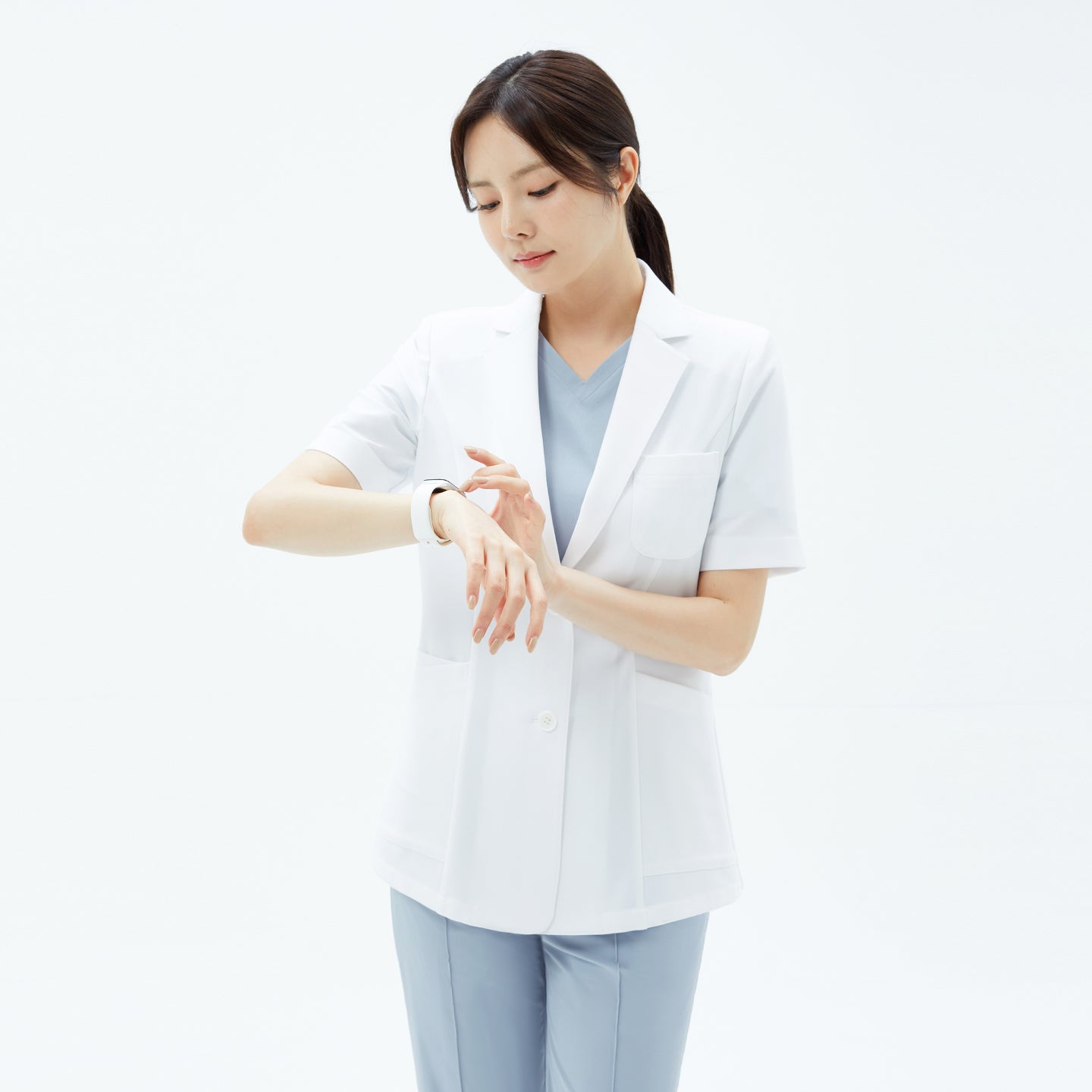 The model in the TUL Women’s LCW-15 Lightweight Short Sleeve Lab Coat – White glances down to tap her smartwatch, highlighting the coat’s breast pocket and structured short sleeves.