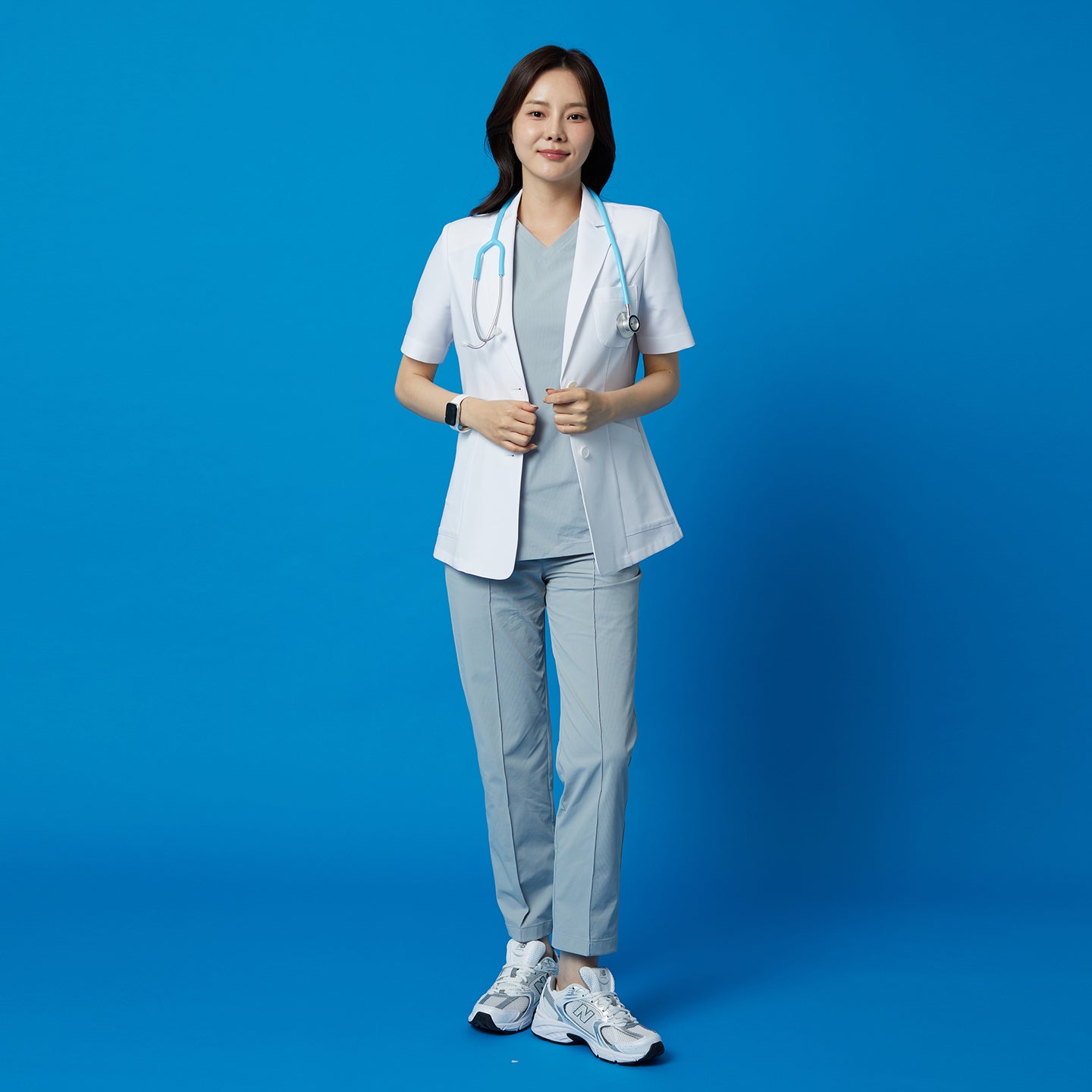Full-length portrait of the TUL Women’s LCW-15 Lightweight Short Sleeve Lab Coat – White worn with a stethoscope around the neck against a solid blue backdrop.