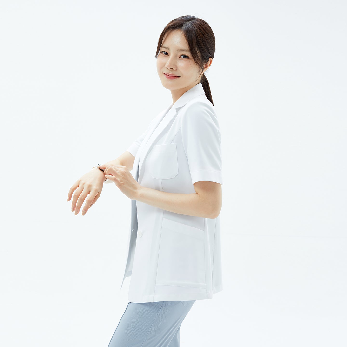 Image of The model in the TUL Women’s LCW-15 Lightweight Short Sleeve Lab Coat – White tapping her smartwatch, highlighting the coat’s breast pocket and structured short sleeves.