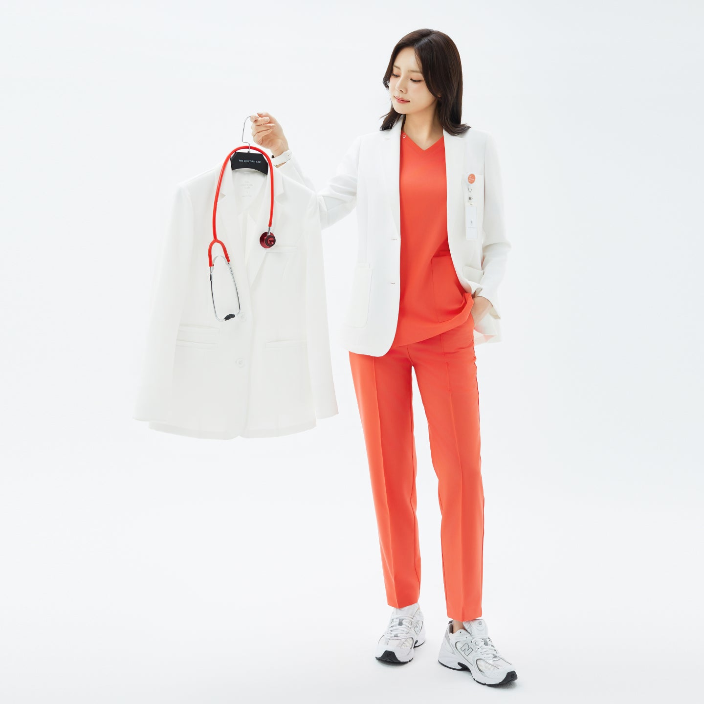 Image of The TUL Women's LCW-16 Prime Short Lab Coat - White Ivory shown in a full-body front view as the model holds another identical coat on a hanger.
