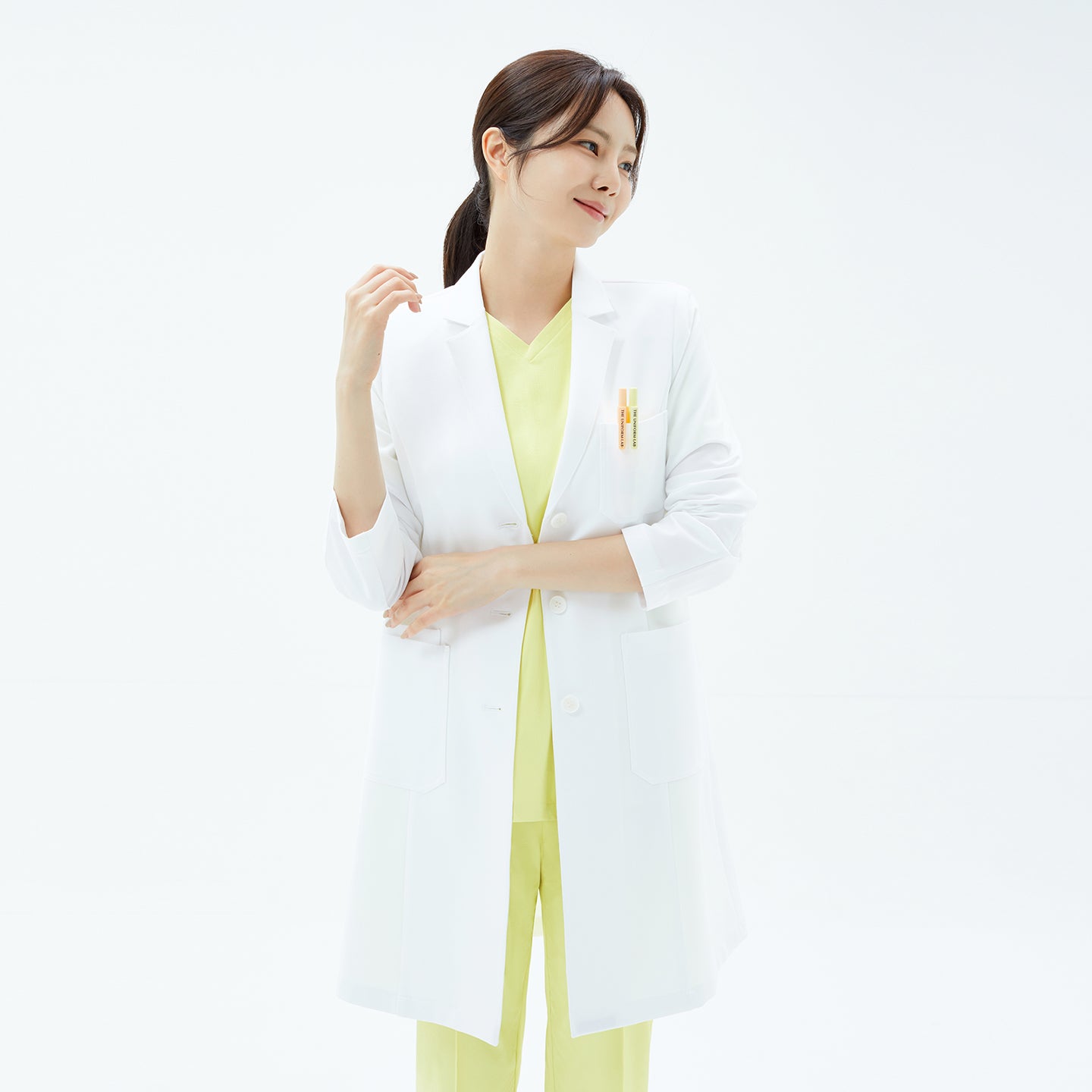 Image of Woman wearing the TUL LCW-17 Straight Long Lab Coat in White over lemon scrubs, crossing arms slightly while facing sideways and smiling.