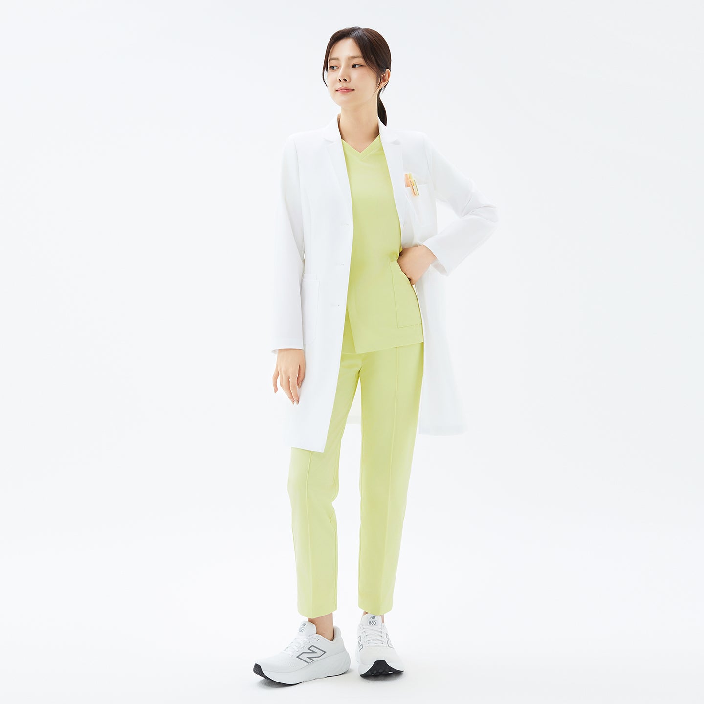Image of Full body shot of the TUL LCW-17 Straight Long Lab Coat in White styled with lemon-colored scrubs and white sneakers.