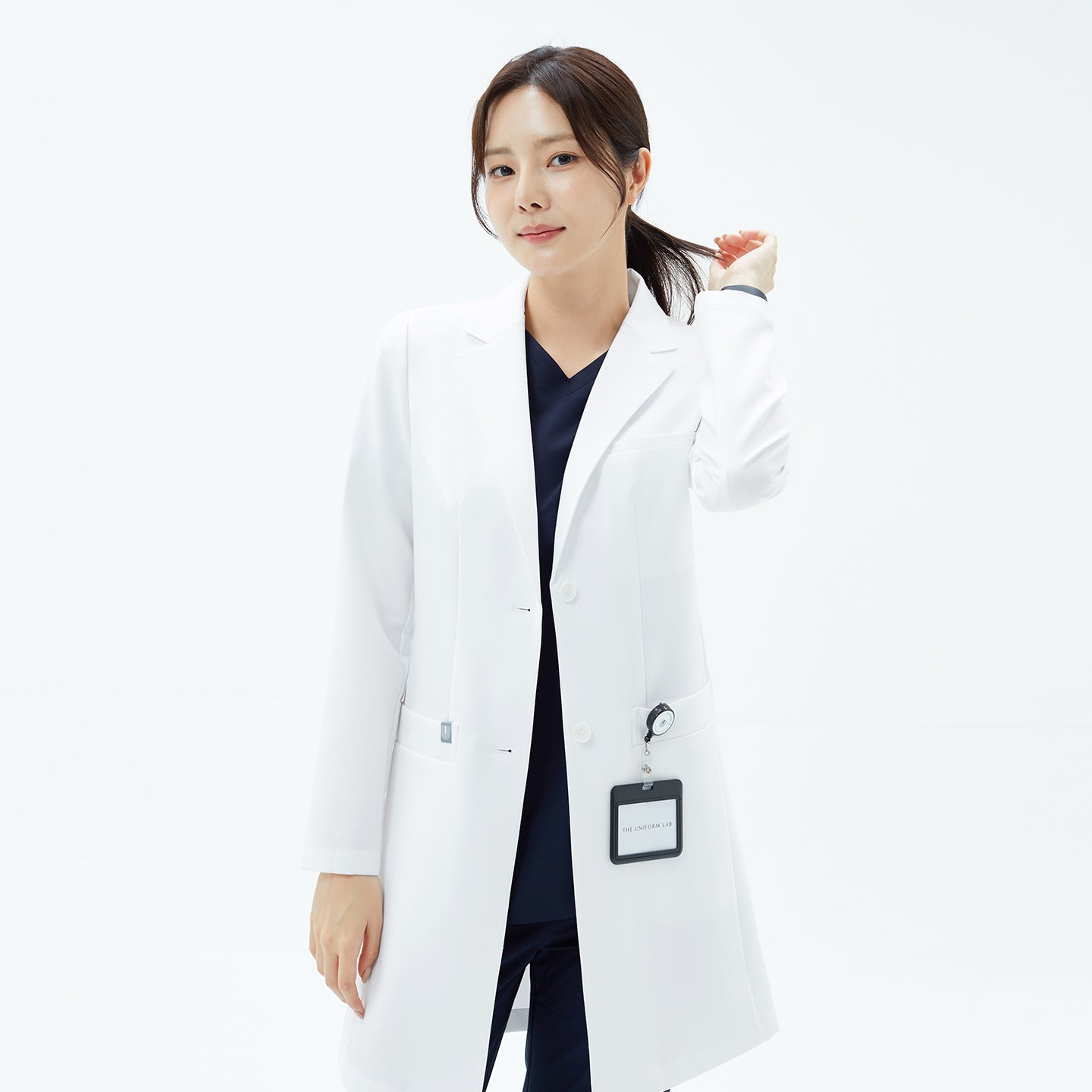 Image of Woman holding ponytail while modeling the TUL Women's LCW-18 Classic Long Lab Coat in White over navy scrubs.