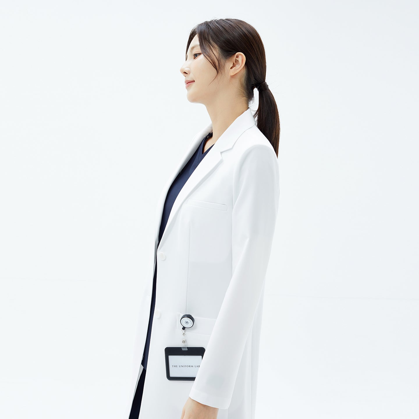 Image of Side profile of woman in the TUL Women's LCW-18 Classic Long Lab Coat in White with stethoscope badge.