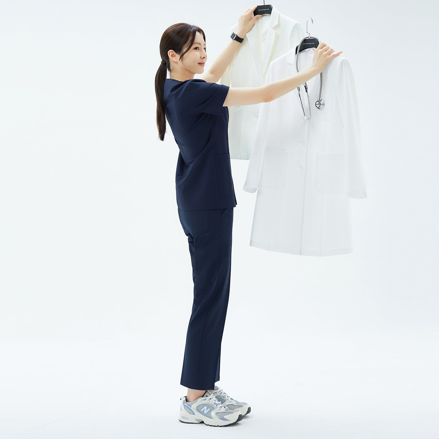 Image of Woman lifting hanger with TUL Women's LCW-18 Classic Long Lab Coat in White next to another lab coat.