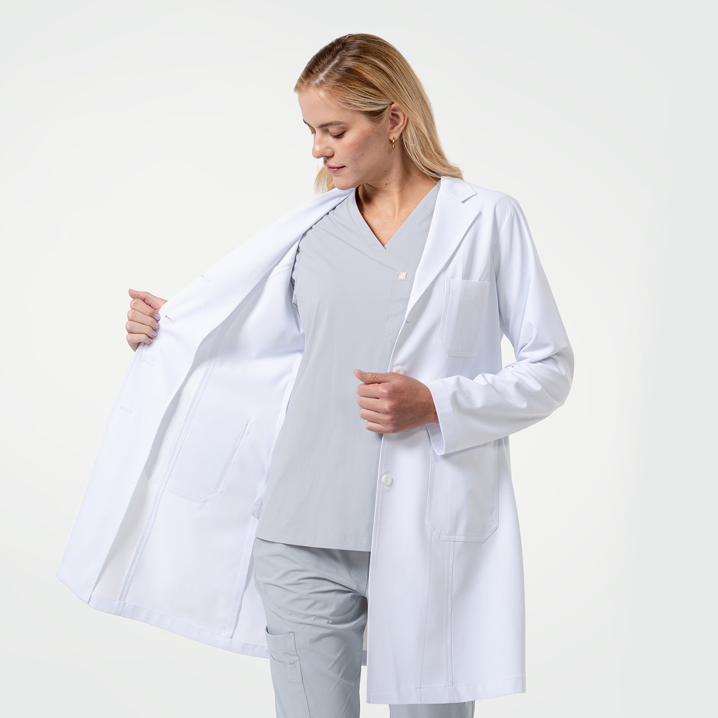 Image of Female doctor wearing a white lab coat over light gray scrubs, looking down while opening the coat to show the inner lining on a light gray background.