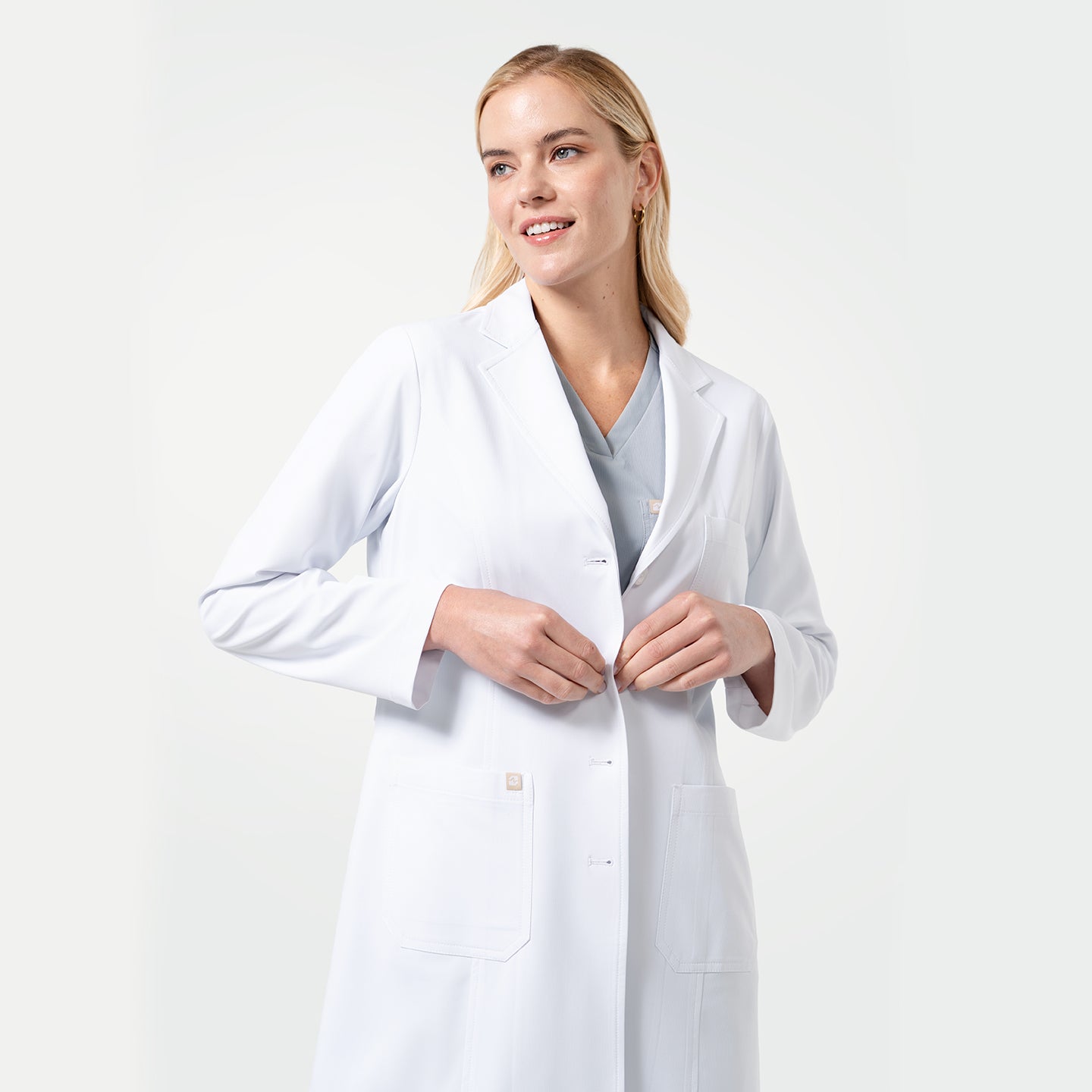 Image of Female model fastening the front buttons of the Women’s Long Lab Coat LCW-04 in white, worn over light gray scrubs.