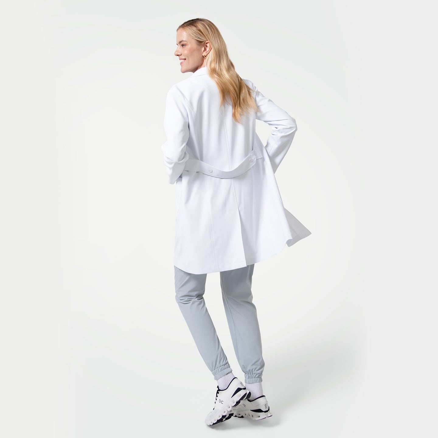 Image of Model facing away from the camera wearing the Women’s Long Lab Coat LCW-04 in white, showing the back belt detail, paired with light gray jogger scrub pants and white sneakers.