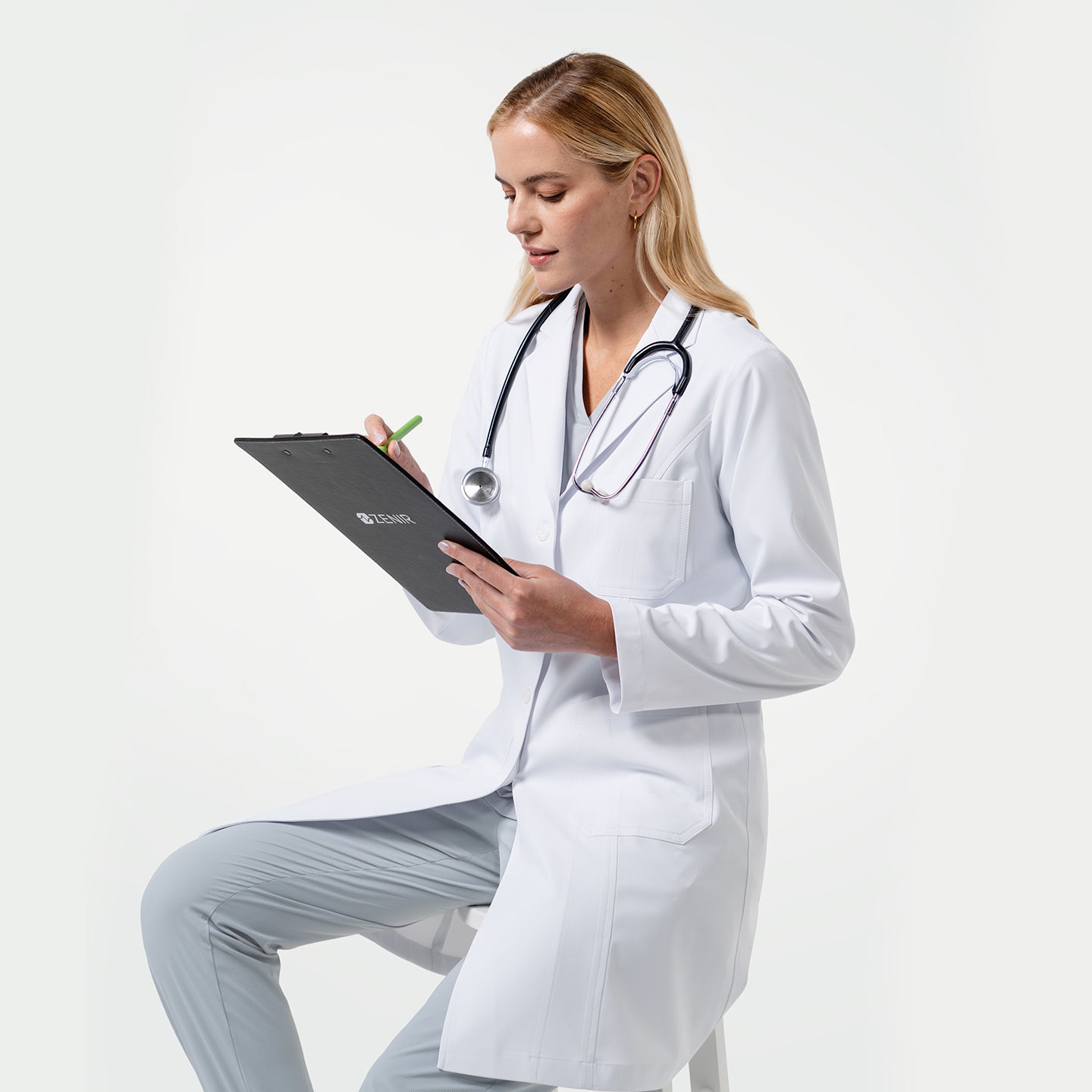 Image of Model wearing a white medical lab coat and stethoscope, sitting on a stool while taking notes on a clipboard against a light gray background.