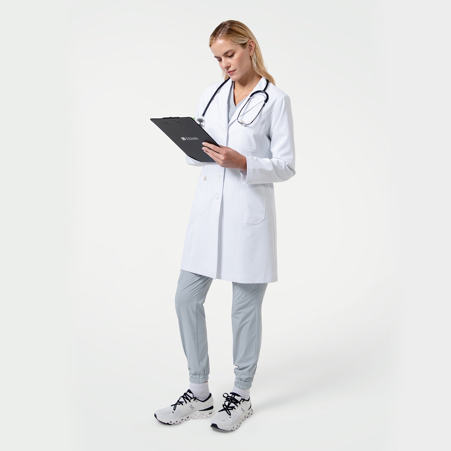 Image of Female doctor wearing a white lab coat with a stethoscope around her neck, holding a clipboard and reviewing notes on a light gray background.