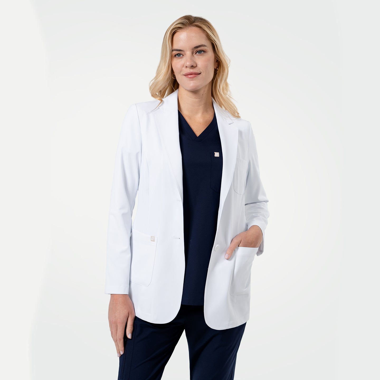 Image of Confident female healthcare professional wearing a white coat layered over navy scrubs, standing with one hand in her pocket on a clean light gray background.