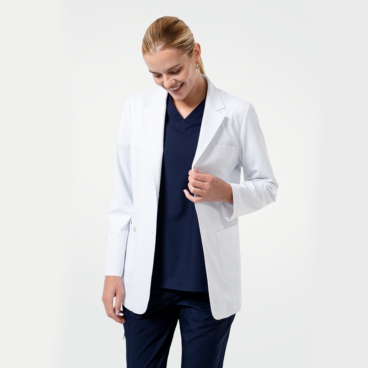 Image of Smiling female healthcare professional wearing a white lab coat over navy scrubs, looking down with a gentle expression on a light gray background.