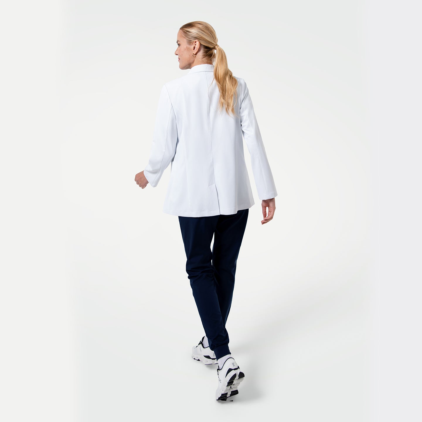 Image of Back view of the Women’s Short Lab Coat LCW-02 in white, showing the model walking away with the coat’s straight hemline and side vents visible, paired with navy scrub pants and white sneakers.