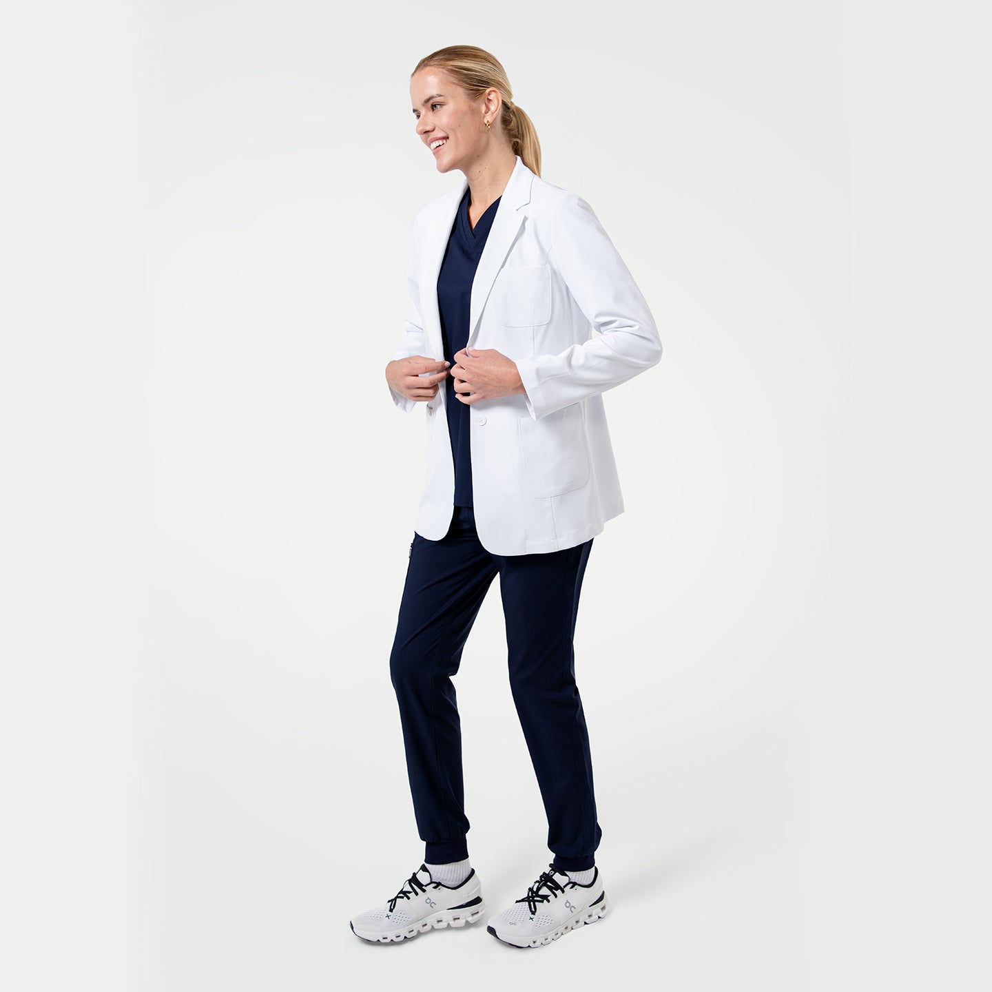 Image of Model wearing a white medical lab coat layered over navy scrubs, smiling and adjusting the front of the coat on a light gray background.