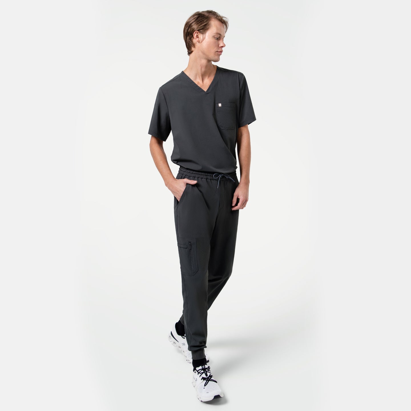 Image of Male model walking with hands in pockets, wearing a matching V-neck scrub top and Zipper Pocket Jogger Scrub Pants in Deep Gray, paired with white sneakers.