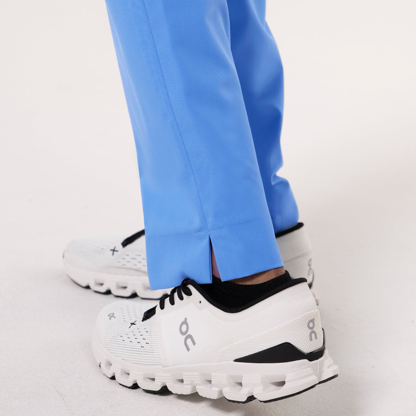 Image of Close-up view of the lower leg section of Ceil Blue zipper pocket split hem scrub pants, showing the small side slit at the hem and white athletic shoes.