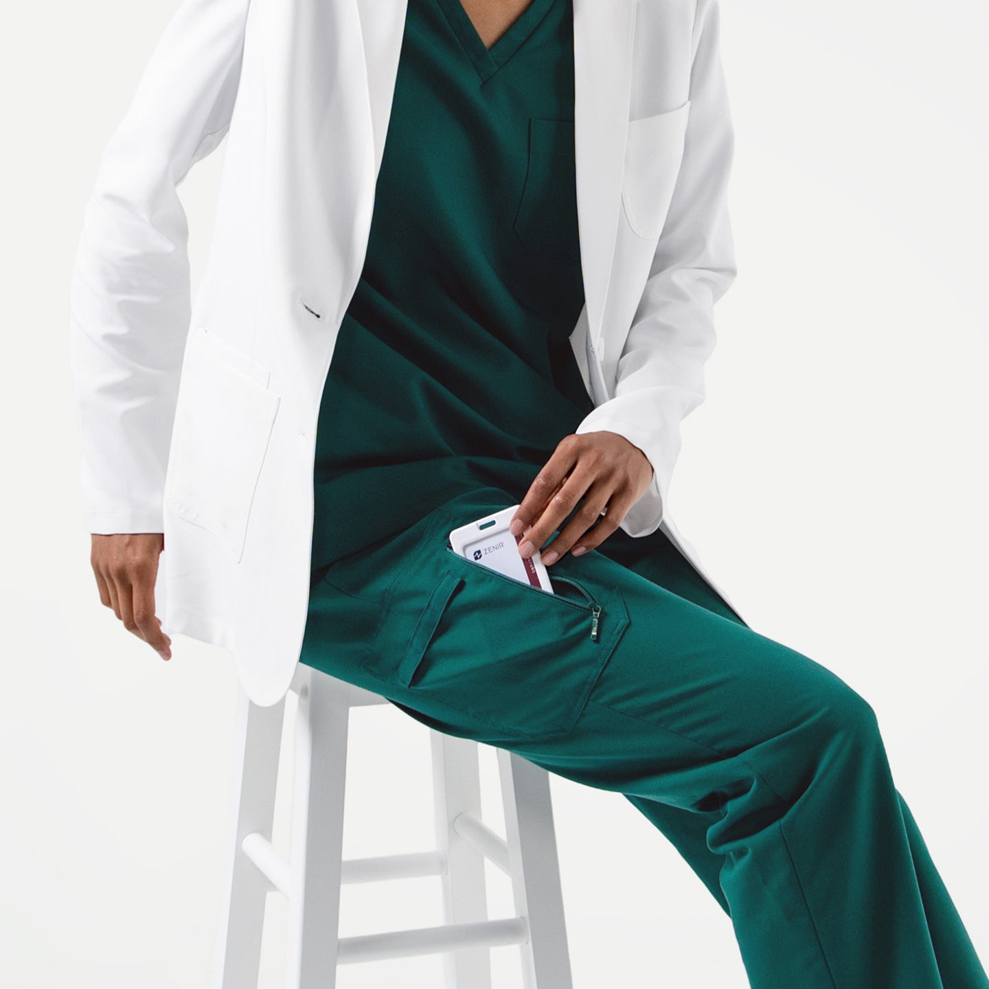 Image of Person wearing a white lab coat over Pine Green zipper pocket scrub pants, seated on a white stool while placing a phone into the pants pocket.