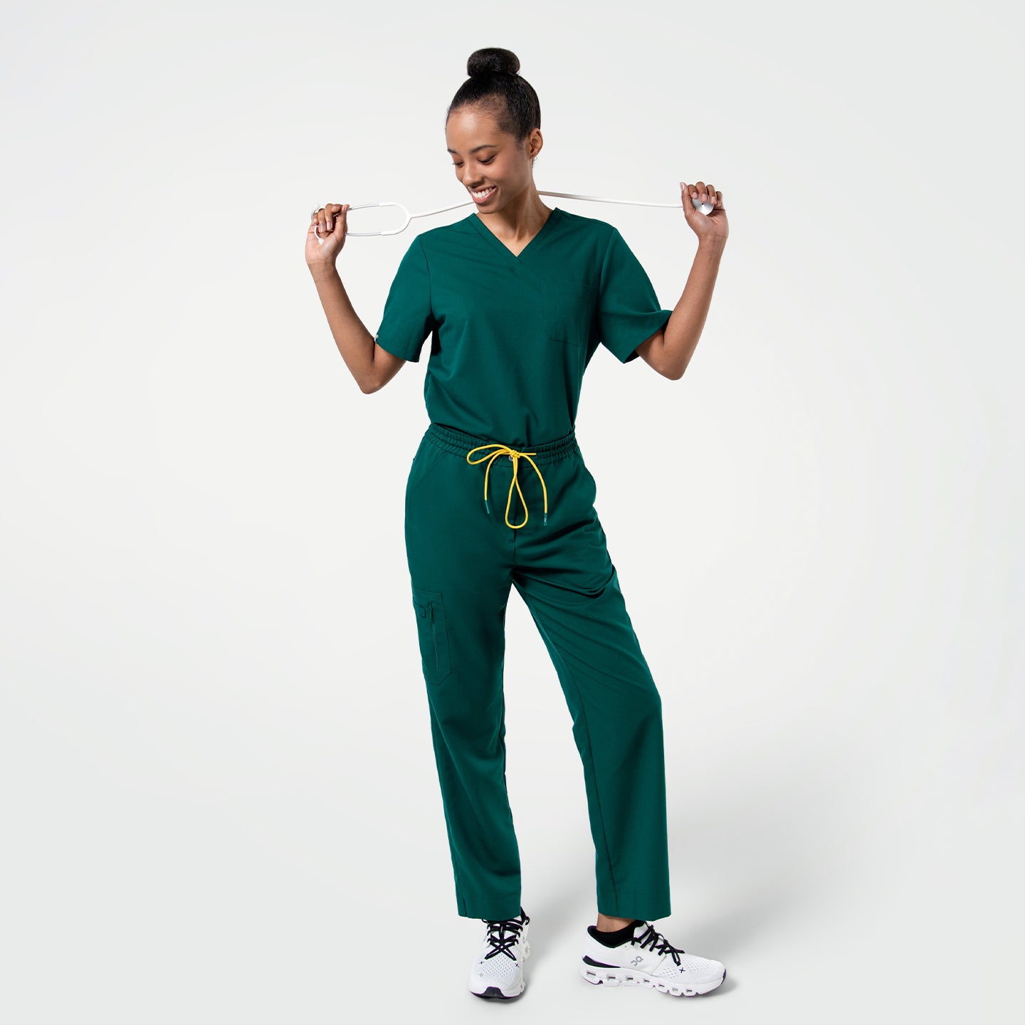 Image of Person wearing Pine Green zipper pocket split hem scrub pants and a matching V-neck scrub top, standing with a stethoscope held playfully across their shoulders.