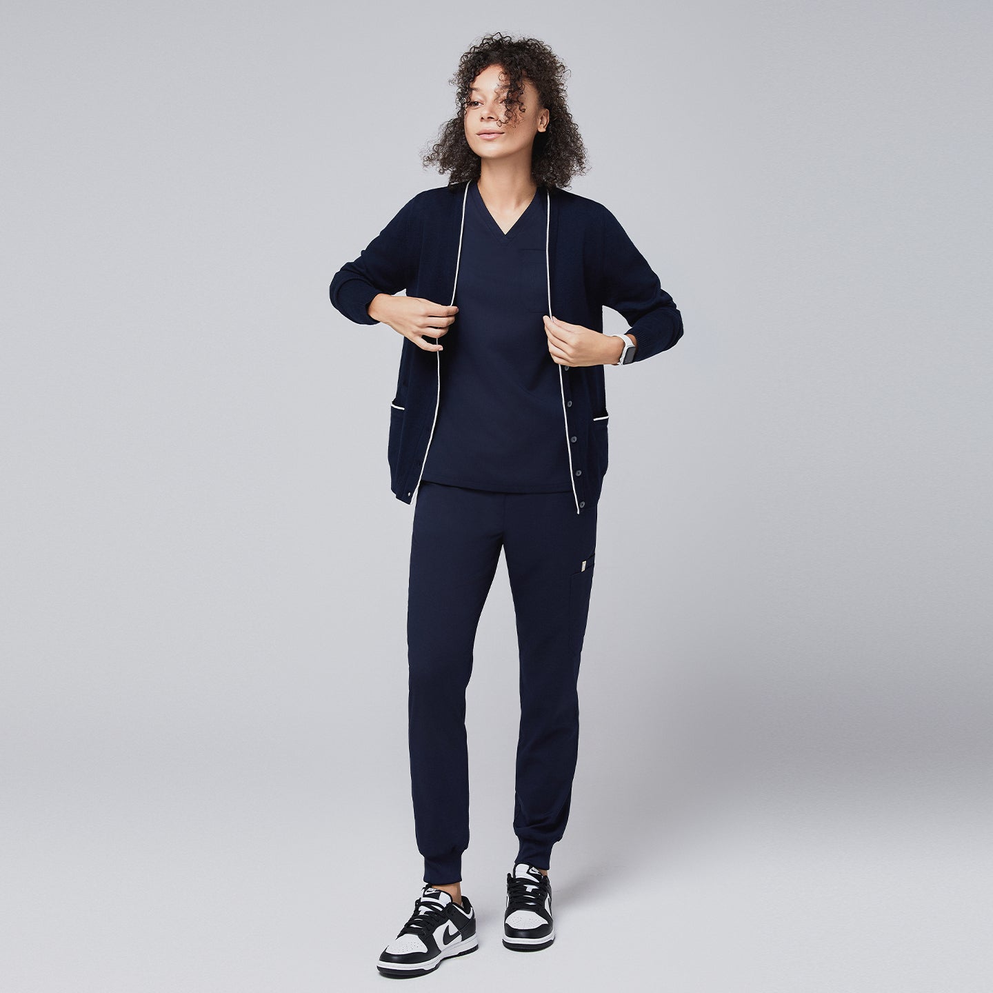 Image of Woman wearing a navy scrub set paired with a matching cardigan, standing confidently with hands adjusting the cardigan, highlighting a professional and comfortable look,Navy